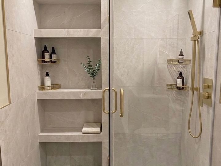 Marble-look tile shower with built-in shelves and gold fixtures behind a glass door - Sani Building Corp